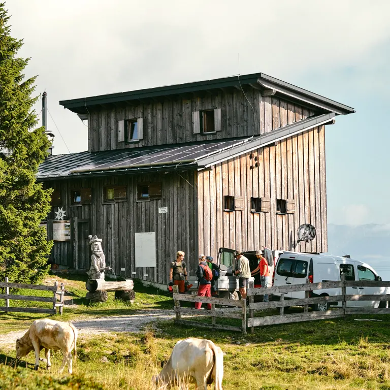 Een groep mensen laadt spullen uit witte bestelbusjes uit bij een houten hut in een landelijk, bergachtig gebied. Op de voorgrond grazen koeien bij een houten hek en naast de hut staat een houten beeld met houtsnijwerk. Een groep mensen laadt spullen uit witte bestelbusjes uit bij een houten hut in een landelijk, bergachtig gebied. Op de voorgrond grazen koeien bij een houten hek en naast de hut staat een houten beeld met houtsnijwerk.